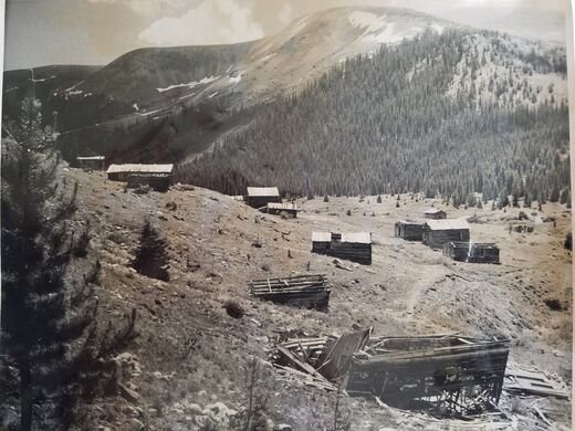 Black and white photograph of a mining camp on a high slope with log cabins and a collapsed timber structure