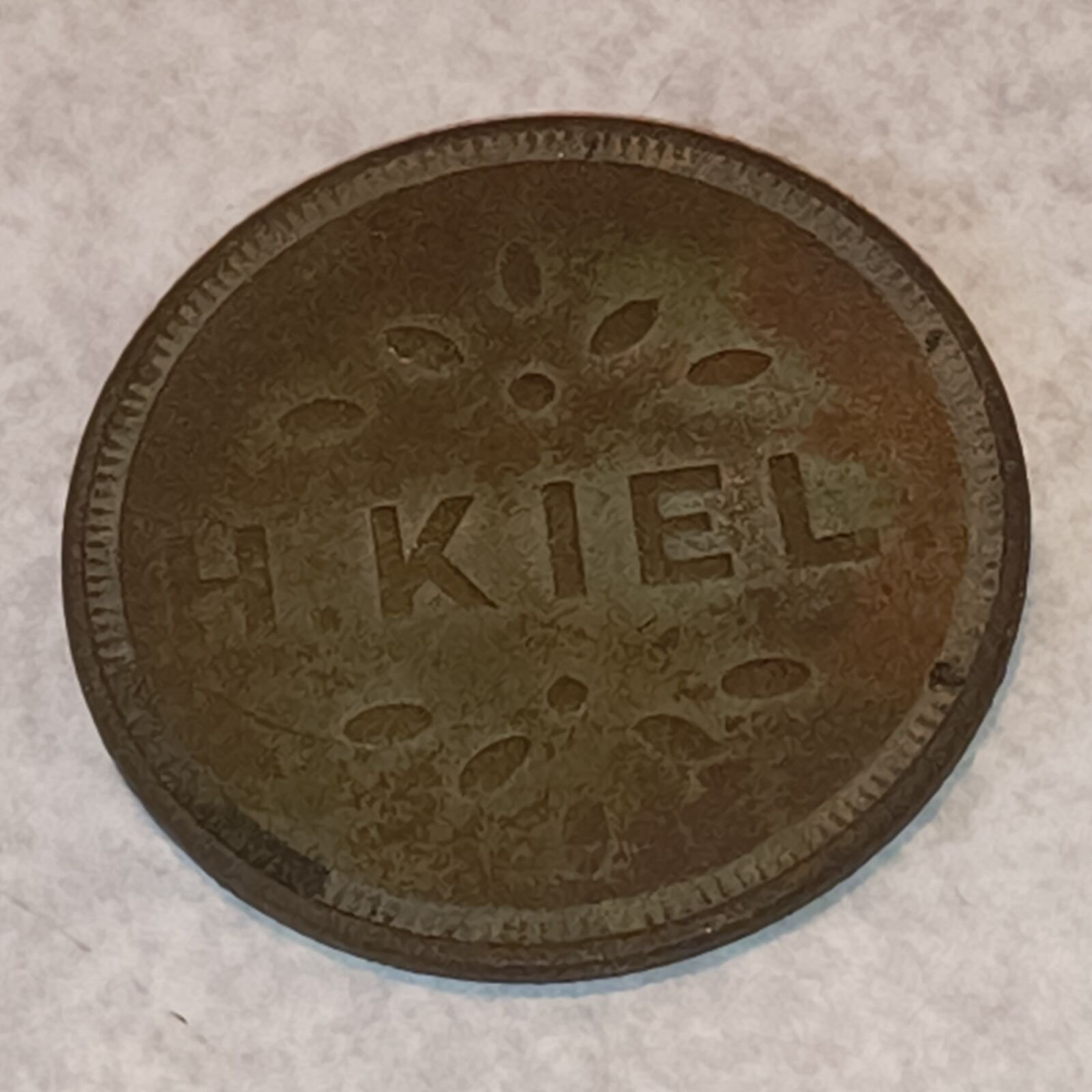 Worn brass token stamped H. KIEL between two sprays of leaves