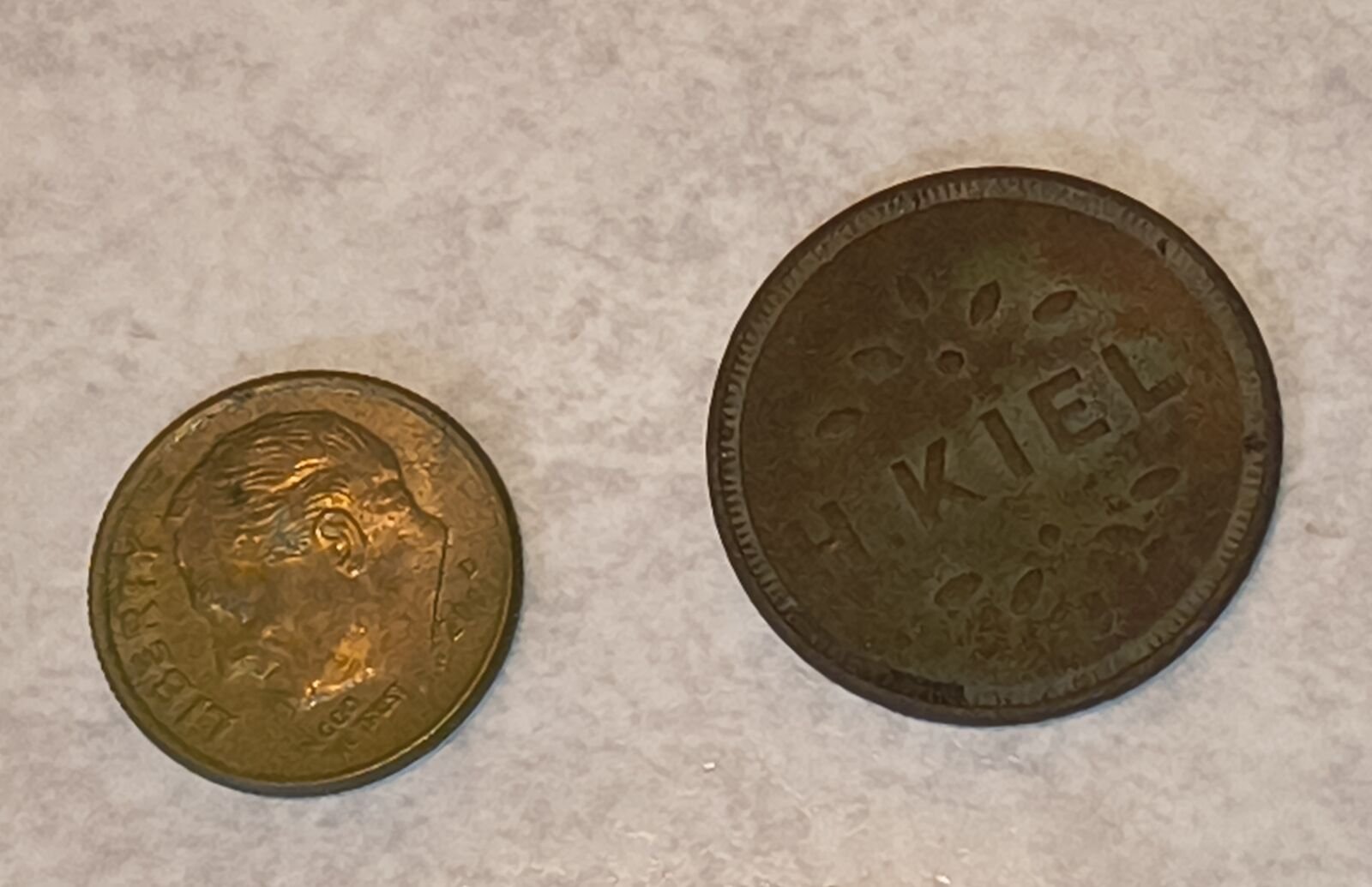 The H. Kiel brass token lying beside a modern United States dime on a white surface