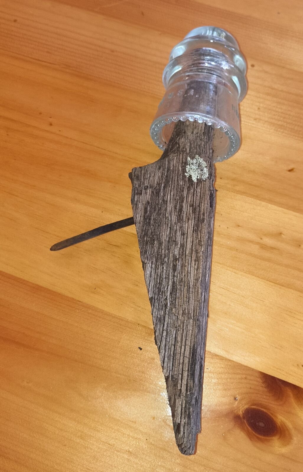 Clear glass insulator threaded onto a weathered wooden pin fragment with an iron spike through it, lying on a wooden table